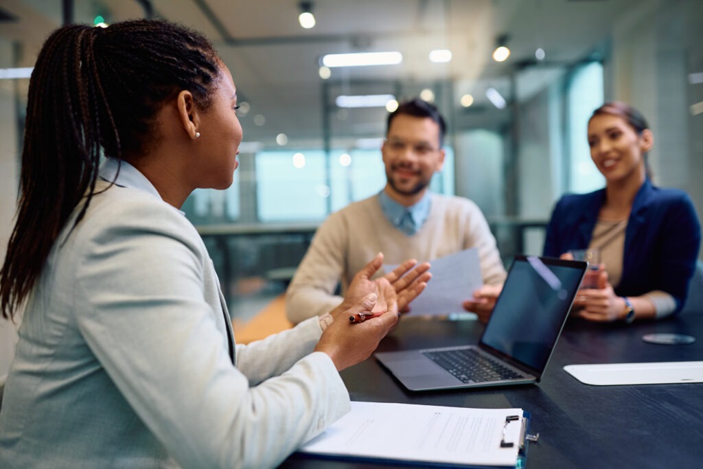 A woman gestures while talking to two colleagues at a conference table. Open laptops and documents suggest a professional meeting. Warm, collaborative tone.